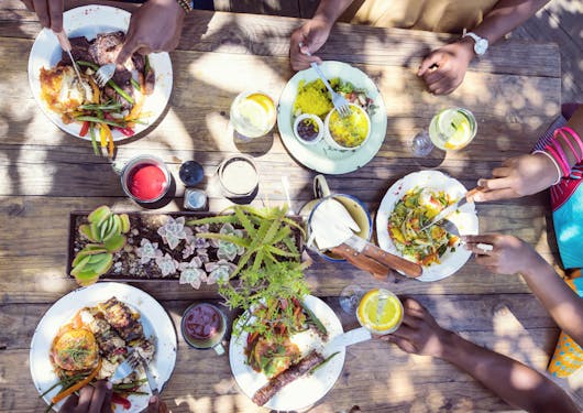 An aerial view shows at least five Black people surrounding a wooden table, each one with a plate of traditional South African food in front of them. They seem to be in a shaded area with sun peeking through leaves.