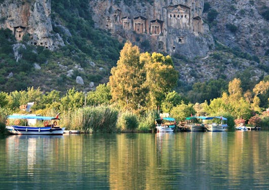 Four small boats with canopies over them sit alongside a shore beneath a large hill with a rocky face and and Roman ruins seemingly built into the face of the rock.
