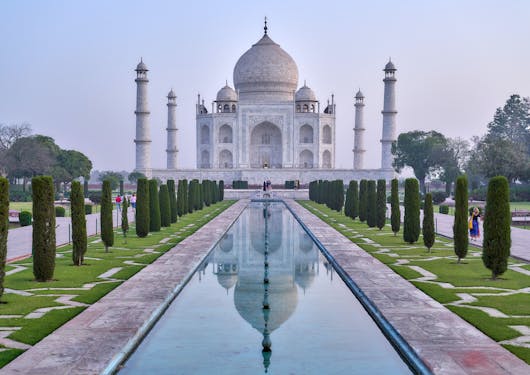 The Taj Mahal is shown at sunrise, the sky is a light hazy purple blue and there are very few people along the tree-lined paths centered in front of the building. There is a still body of water in a rectangular form straight in front of the Taj Mahal.