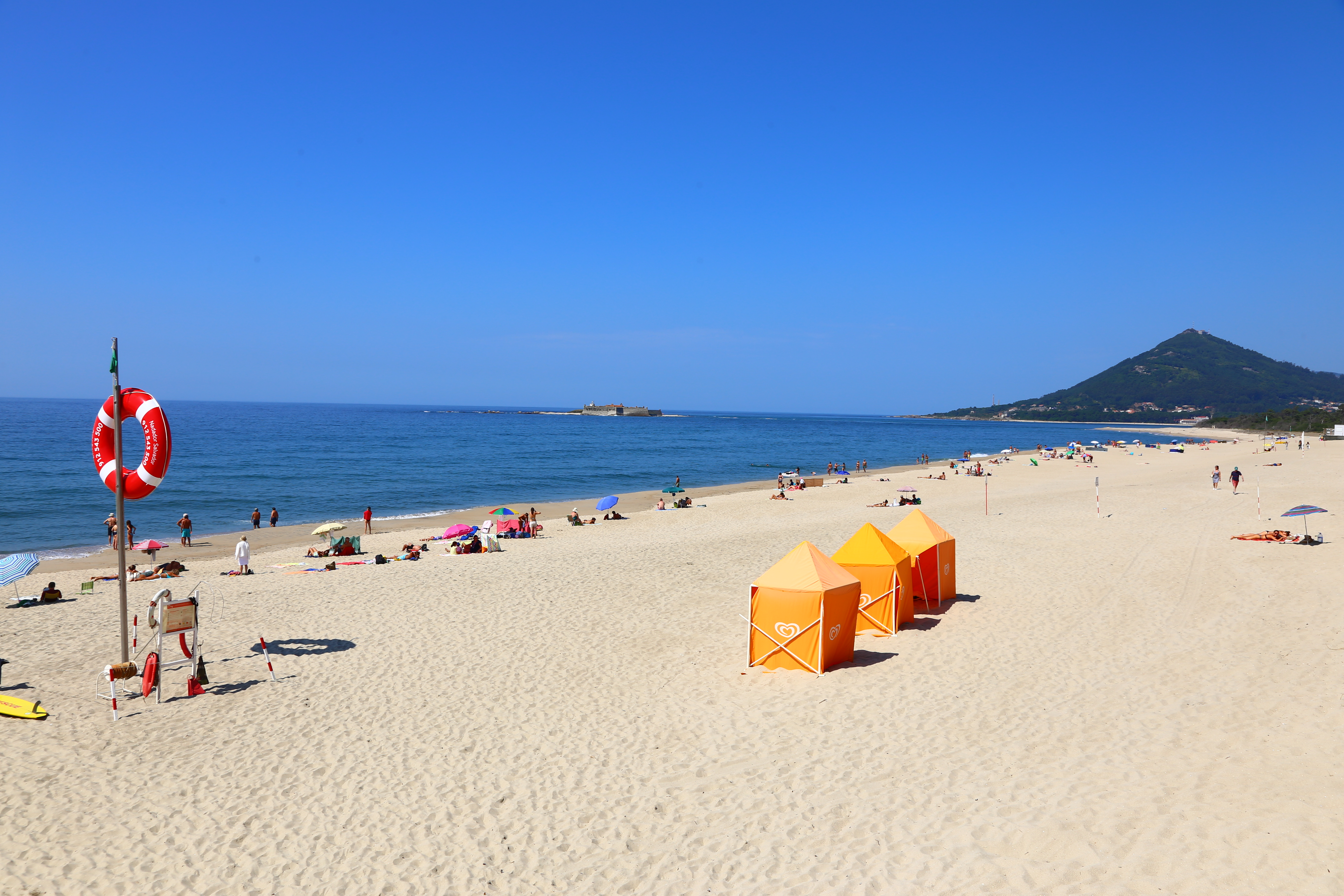A white sand beach with blue ocean to the left and blue sky above has 3 orange beach tents on it, and sun bathers near the shore.