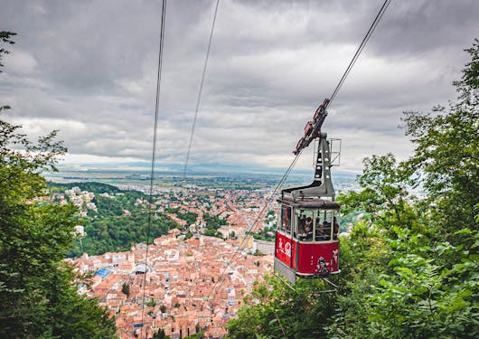 The city of Brasov is shown from up above as a funicular or cable car comes up the mountain. There are green trees on either side of the frame and it's a cloudy day.