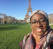 A headshot of a young Black woman wearing a head band, scarf, and jacket, standing in front of the Eiffel Tower on the green grass of the Champs de Mars on a blue-sky sunny day.