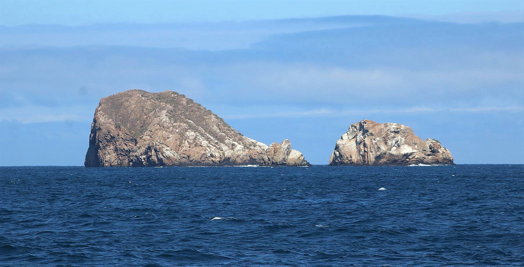 Two islands, one larger on the left and one smaller on the right, are shown in the ocean of the Galapagos Islands in Ecuador. They look very rugged and uninhabitable with steep rocky faces. The sky is blue on a sunny day.