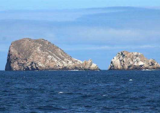 Two islands, one larger on the left and one smaller on the right, are shown in the ocean of the Galapagos Islands in Ecuador. They look very rugged and uninhabitable with steep rocky faces. The sky is blue on a sunny day.