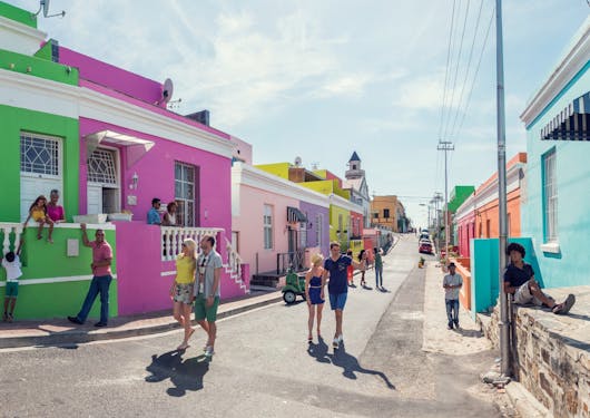 A street lined with delightfully colored houses: green, magenta, pink, purple, yellow, blue, and orange, is shown with local people sitting along the side and walking up the street toward the camera on a sunny day. This is in South Africa.