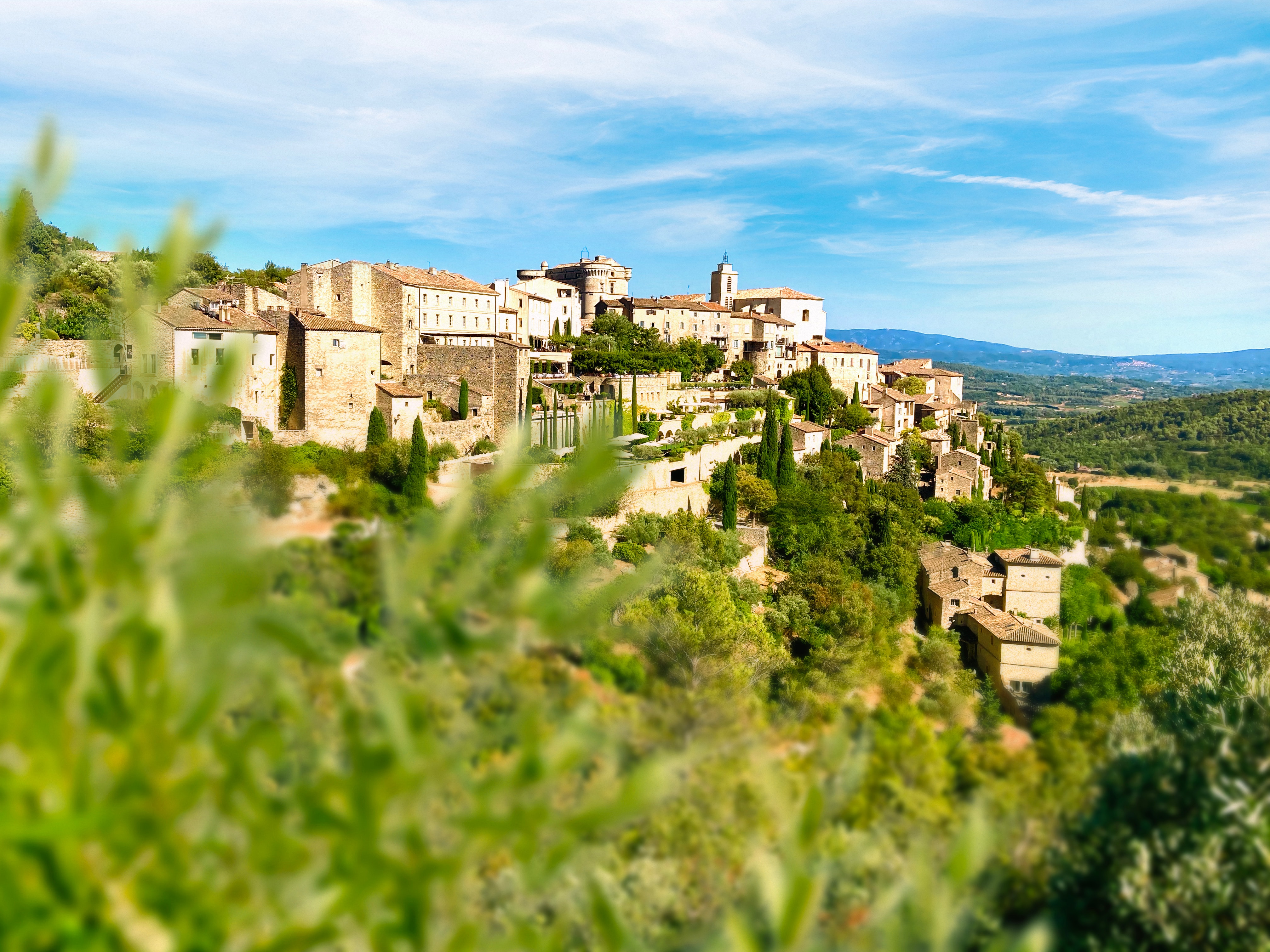 Gordes sits perched in the Luberon region of the French countryside, with green foliage unfocused in the foreground.