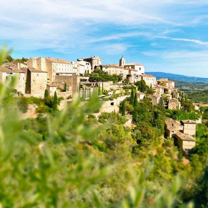 Gordes sits perched in the Luberon region of the French countryside, with green foliage unfocused in the foreground.