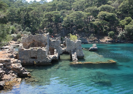 Clear, turquoise water is shallow enough where you can see sunken roman ruins below, with a rocky shore to the left and green trees in the background.