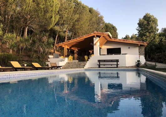 A pool sits at dusk at a yoga retreat in Portugal, with green trees lit by golden hour an the main sustainable retreat center building in the background.