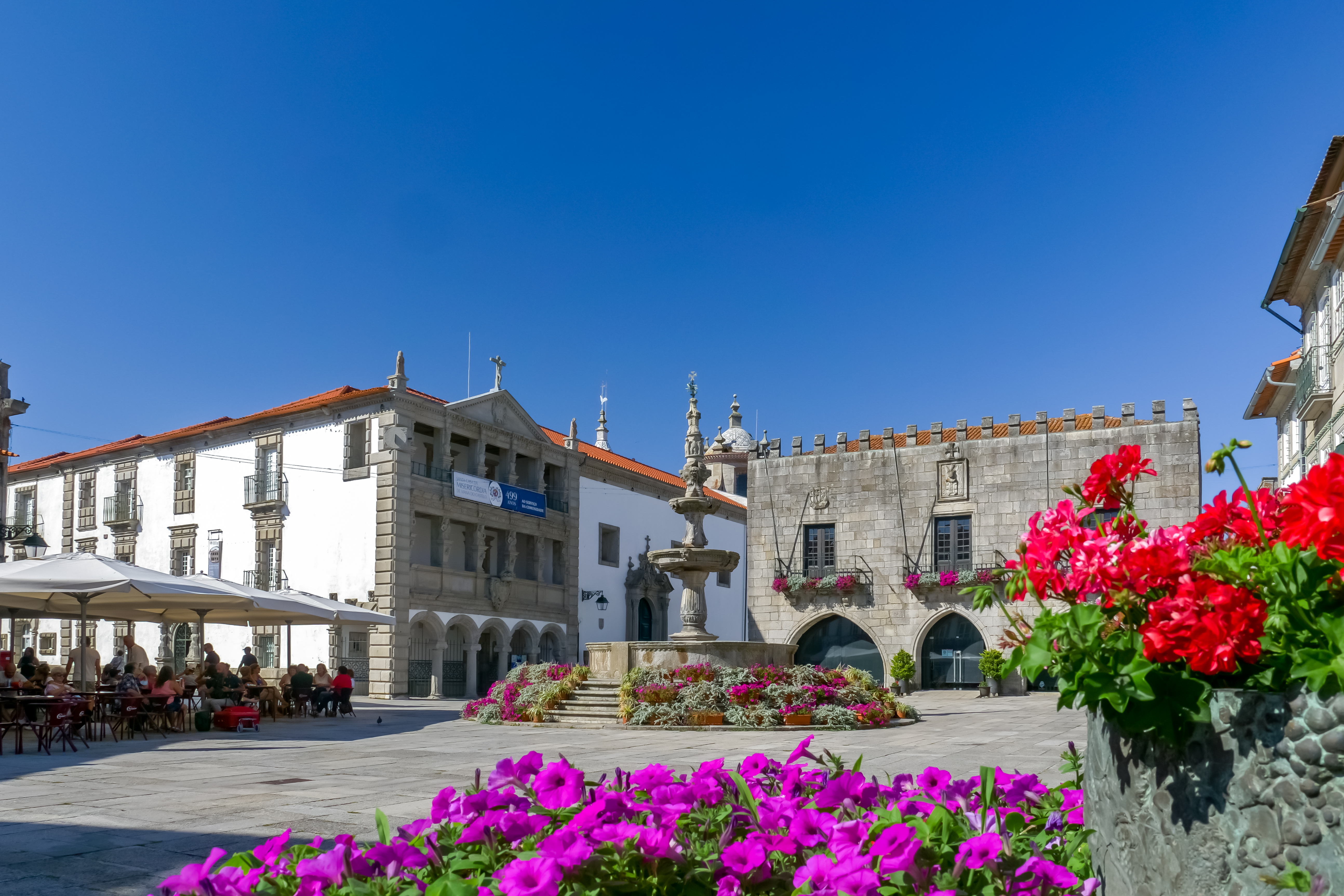 A charming village in northern Portugal is shown in the sunshine with a pure blue sky, purple and red flowers in the foreground, a fountain in the middle, and a covered, terraced cafe to the left. 