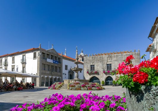 A charming village in northern Portugal is shown in the sunshine with a pure blue sky, purple and red flowers in the foreground, a fountain in the middle, and a covered, terraced cafe to the left.