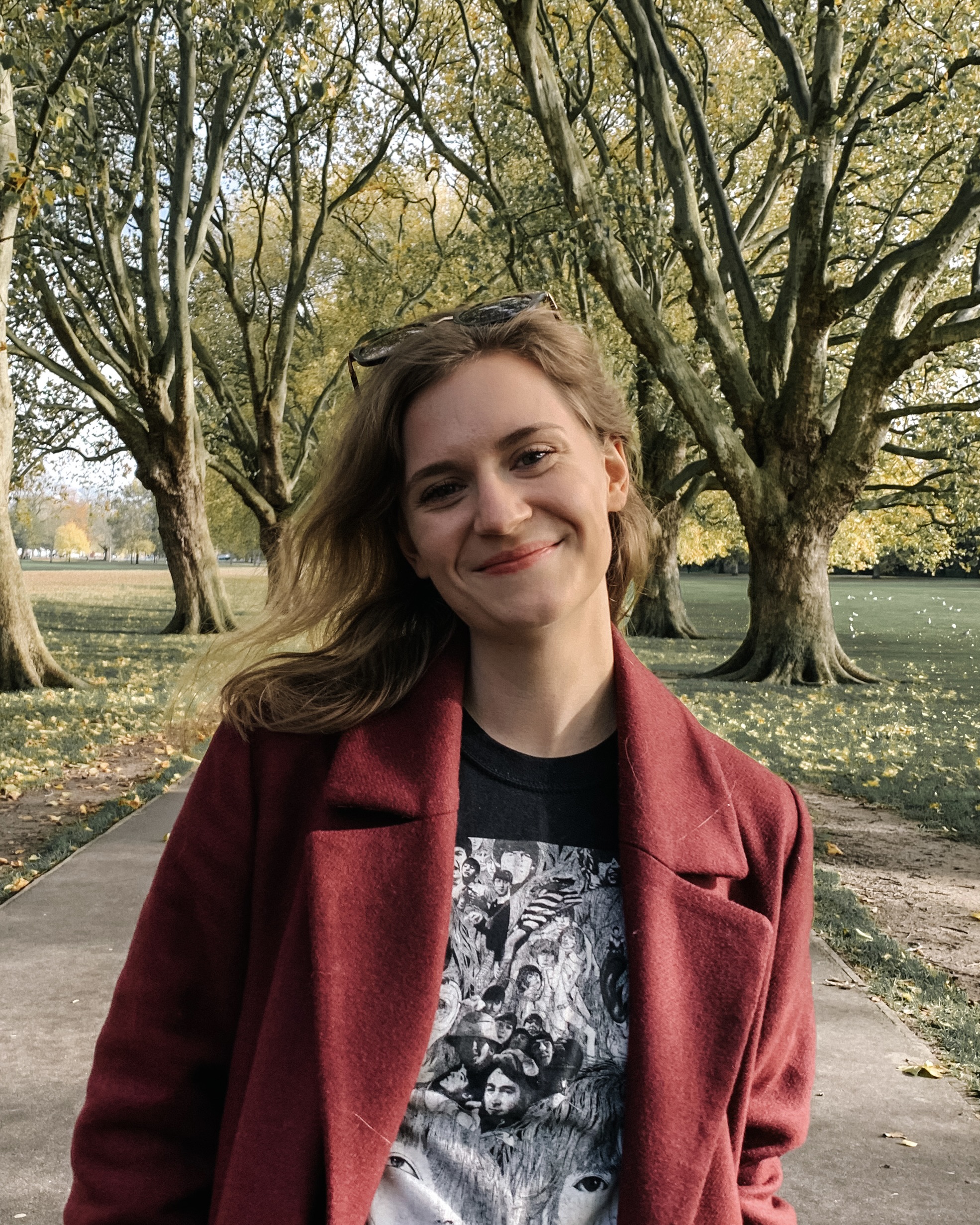 Young Caucasian women stands on a path, smiling, with sunlit trees behind her. She's wearing a red coat over a black screen print t shirt, and has long brown hair worn down.