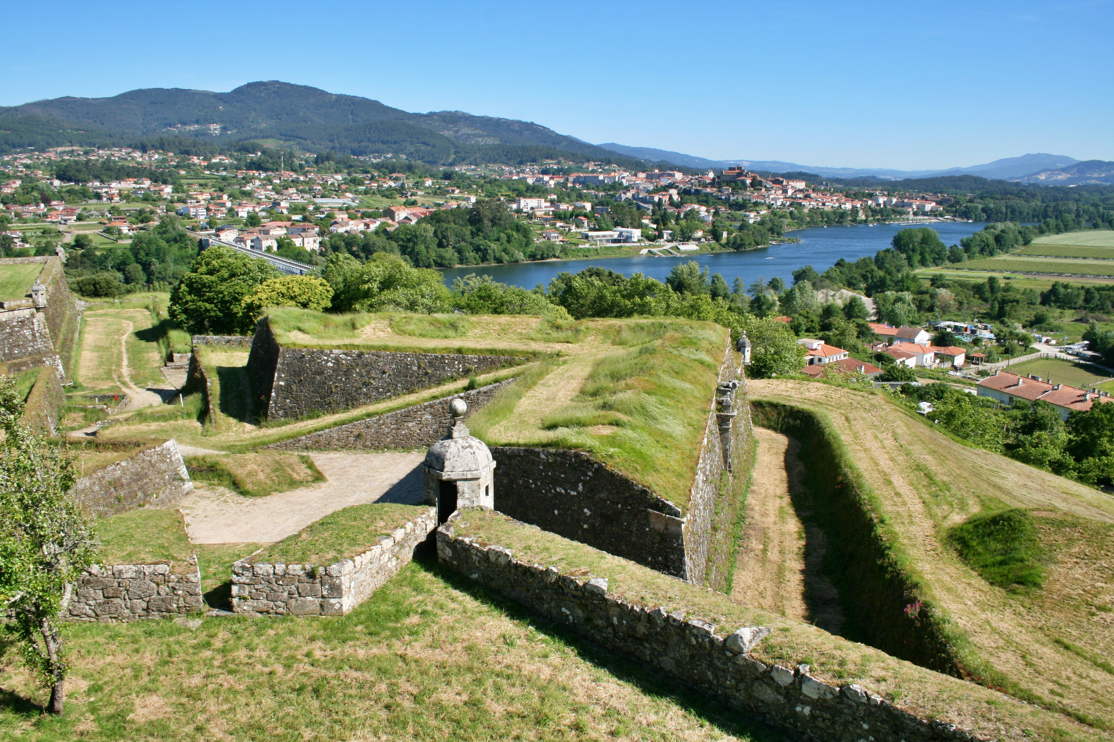 Old, moss-covered fortifications look over a bright blue river and a small town with red-topped houses on the other side of it. It's a sunny day and the sky is blue.