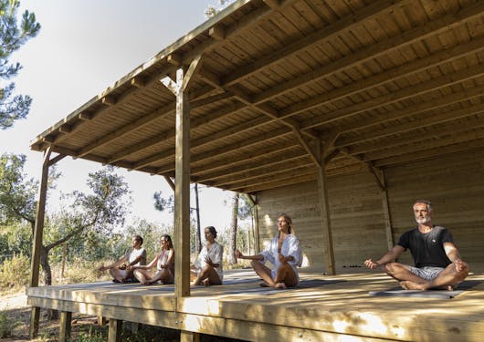 A group of 5 adult yogis sit in a meditative pose with their legs crossed and their arms gently resting on their legs, with their eyes closed. They are in the outdoor wooden open air shala in Portugal at a sustainable yoga retreat.