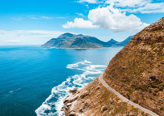 A stunning aerial view of the South Africa coast is show, with bright blue water lapping into a steep brown hillside. A two land road winds around the hill and coastline with a couple of cars on it. More land is in the background, though it's unclear if it's an island or connected the land in the front of the frame. The sky is blue with some white clouds.