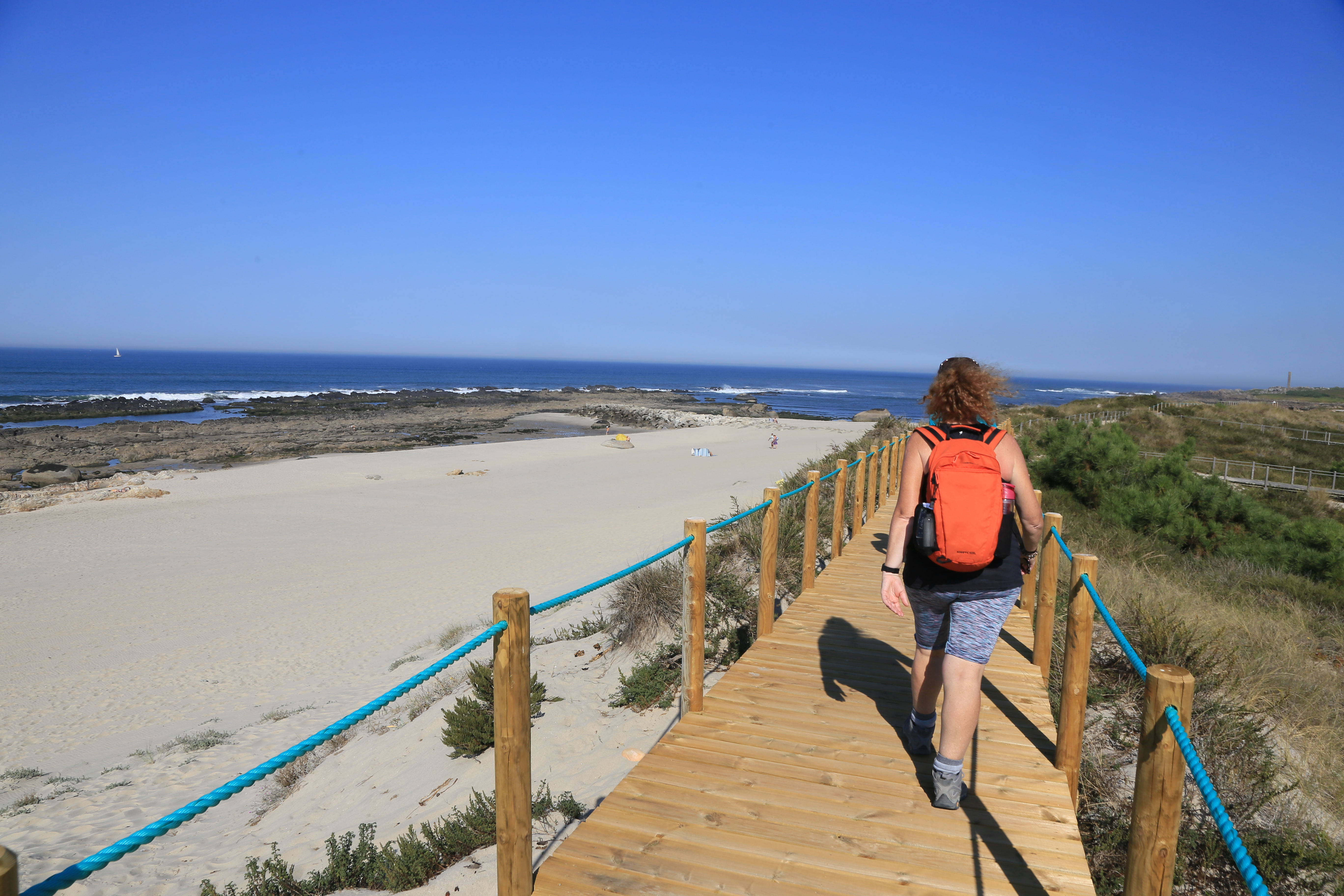 A Causcasian adult woman walks along a boardwalk along Portugal's Atlantic Coast, with sand and ocean to her left.