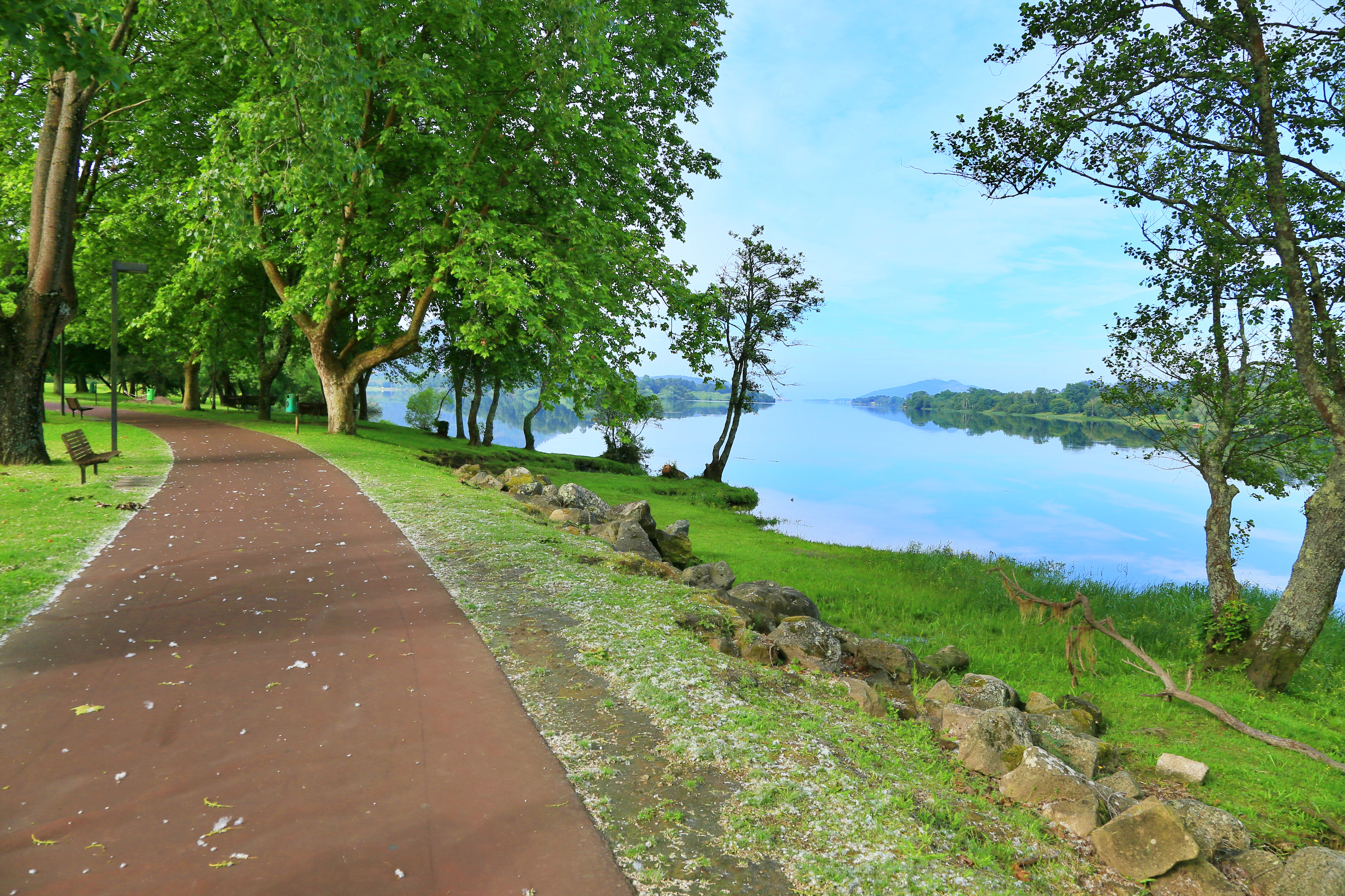 A brownish path for cycling and walking heads into the distance and curves to the left; lush green trees surround the path while a blue river flows to the right.