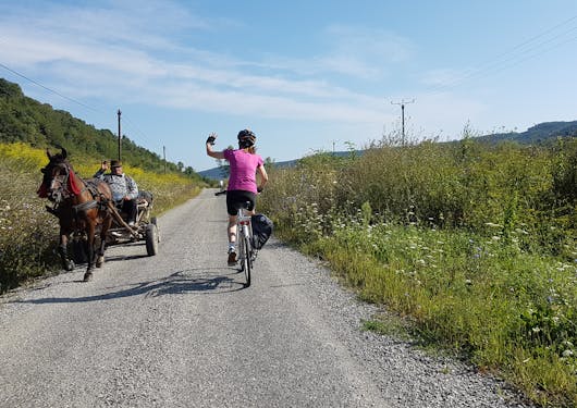 An adult Caucasian female biker wearing black shorts and a magenta top and black helmet bikes on a loose gravel road past a brown horse pulling a cart with a local Romanian man in it. There is greenery and trees on both sides and it's a sunny and partly cloudy day. The woman waves to the man as she passes him; her back is to us and the horse and cart are coming toward the frame.