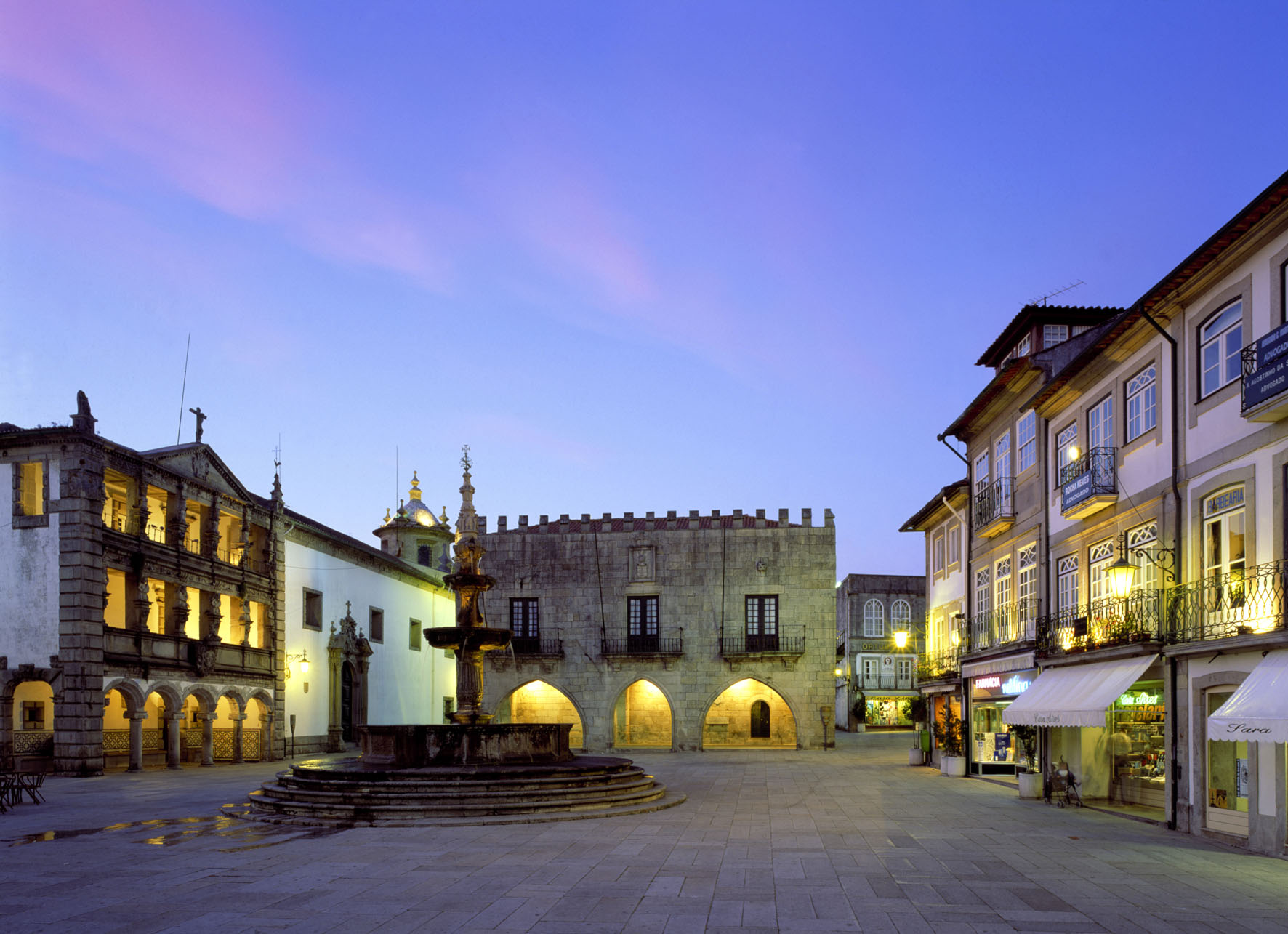 A town in northern Portugal at dusk. A quaint square lined with charming 3-story buildings is under a pink and purple-hued evening sky. There is a fountain in the middle. Buildings are lit up with warm yellow lighting. The square is empty of people.
