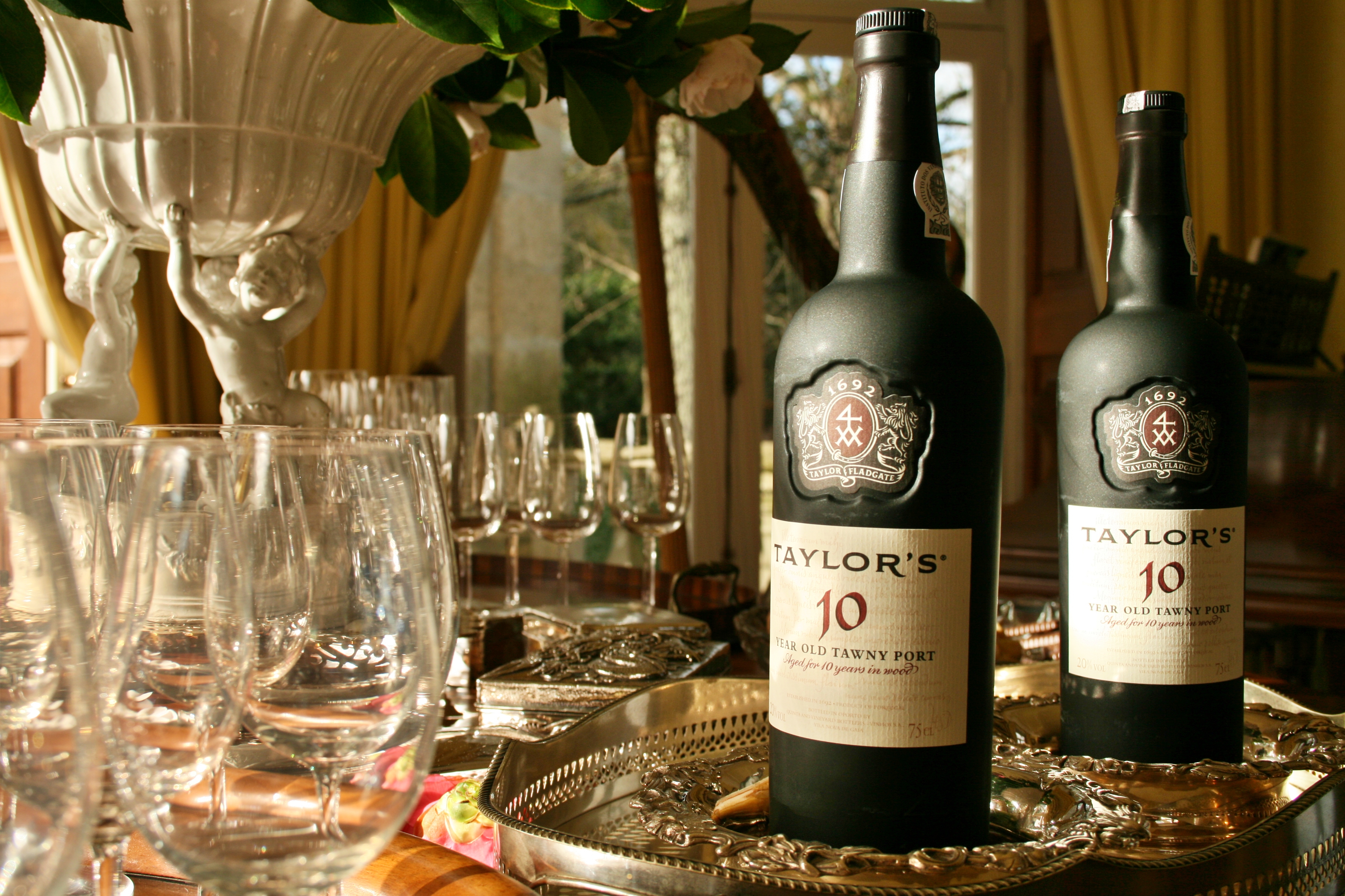 Two black pottles of Tawny Port wine sit on a silver platter off to the right, empty wine glasses are to the left in both the foreground and background. It is staged for a port wine tasting in Portugal.