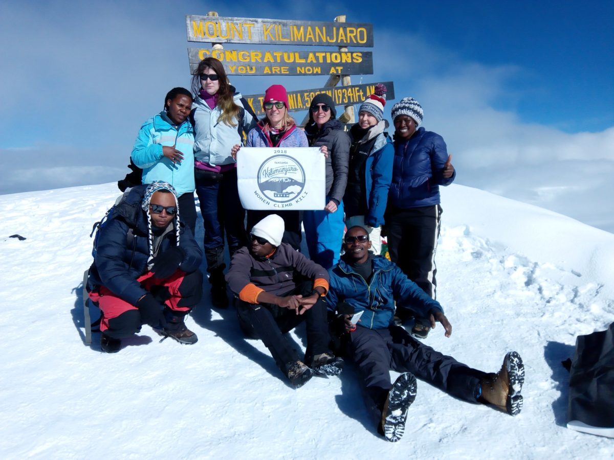 A group of 6 women (2 are Black, on the far left and right, with 4 Caucasian women in the middle) stand atop a snow-capped Mount Kilimanjaro in Tanzania, with three Black men sitting in the front. They are holding a sign that reads "Women Climb Kili" and they are in front of the "Mount Kilimanjaro: Congratulations" sign post - the text is bright yellow. It's a sunny and cloudy day.