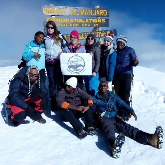 A group of 6 women (2 are Black, on the far left and right, with 4 Caucasian women in the middle) stand atop a snow-capped Mount Kilimanjaro in Tanzania, with three Black men sitting in the front. They are holding a sign that reads "Women Climb Kili" and they are in front of the "Mount Kilimanjaro: Congratulations" sign post - the text is bright yellow. It's a sunny and cloudy day.