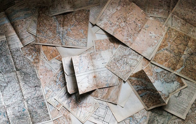 The entire image area is covered with overlapping paper maps in mostly sepia tones, they are open after having been folded; you can see the creases. The maps look well-loved.