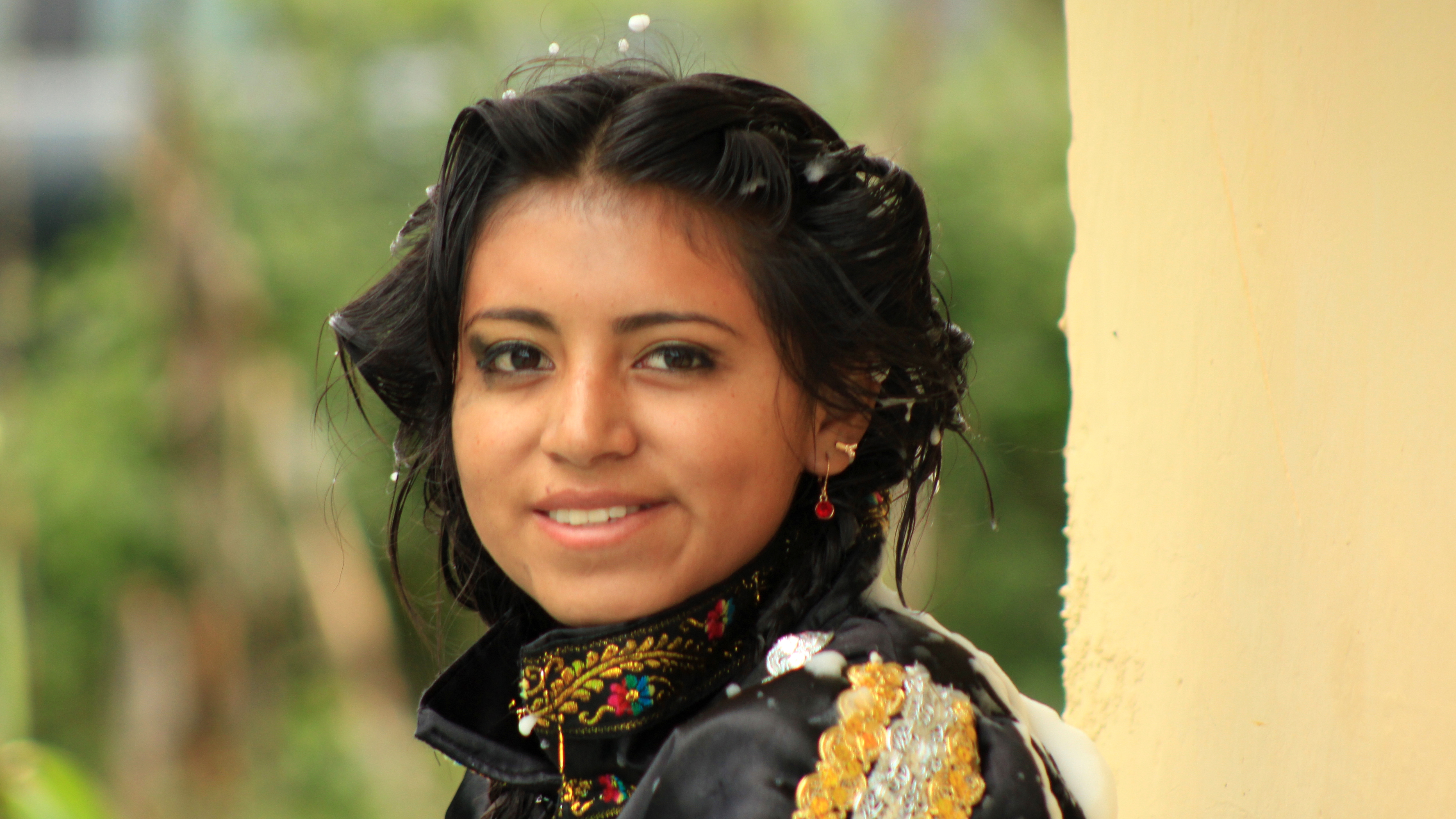 A local Ecuadorian woman in traditional dress (black clothes embellished with gold and colors) smiles warmly at the camera. The green and beige background is blurred.