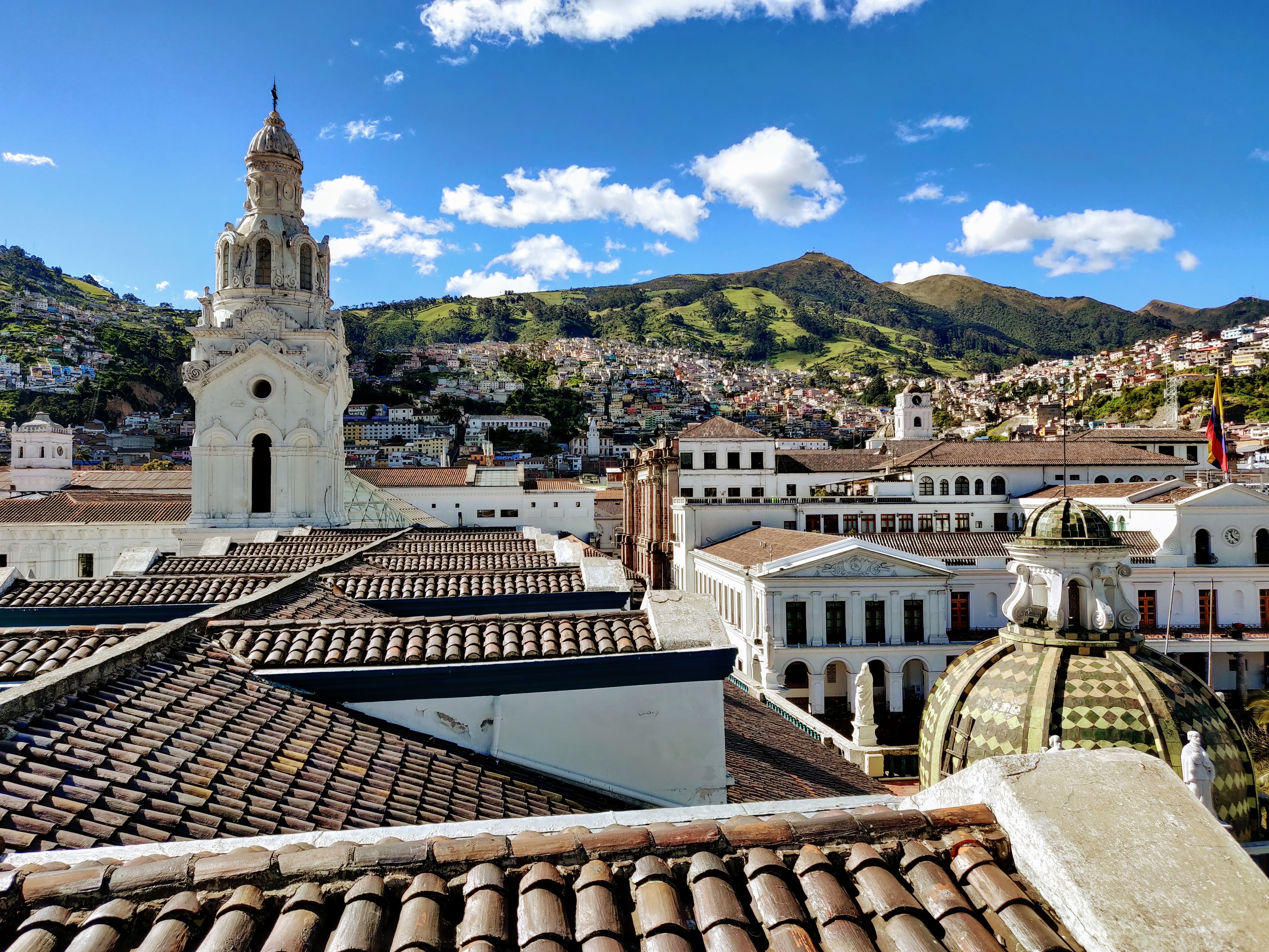 A view from above in Quito, Ecuador shows rooftops in the foreground, as well as the top of a cathedral, with green hills in the background under a  blue sky.