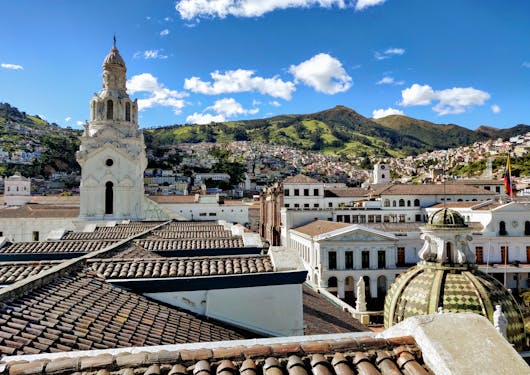 A view from above in Quito, Ecuador shows rooftops in the foreground, as well as the top of a cathedral, with green hills in the background under a blue sky.