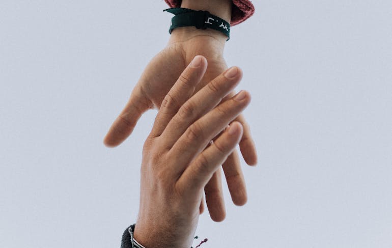 Two hands extend toward each other to grab one another, one from the bottom of the frame and the other from the top of the frame. Both are wearing dark long sleeves and some bracelets. The hands are white.