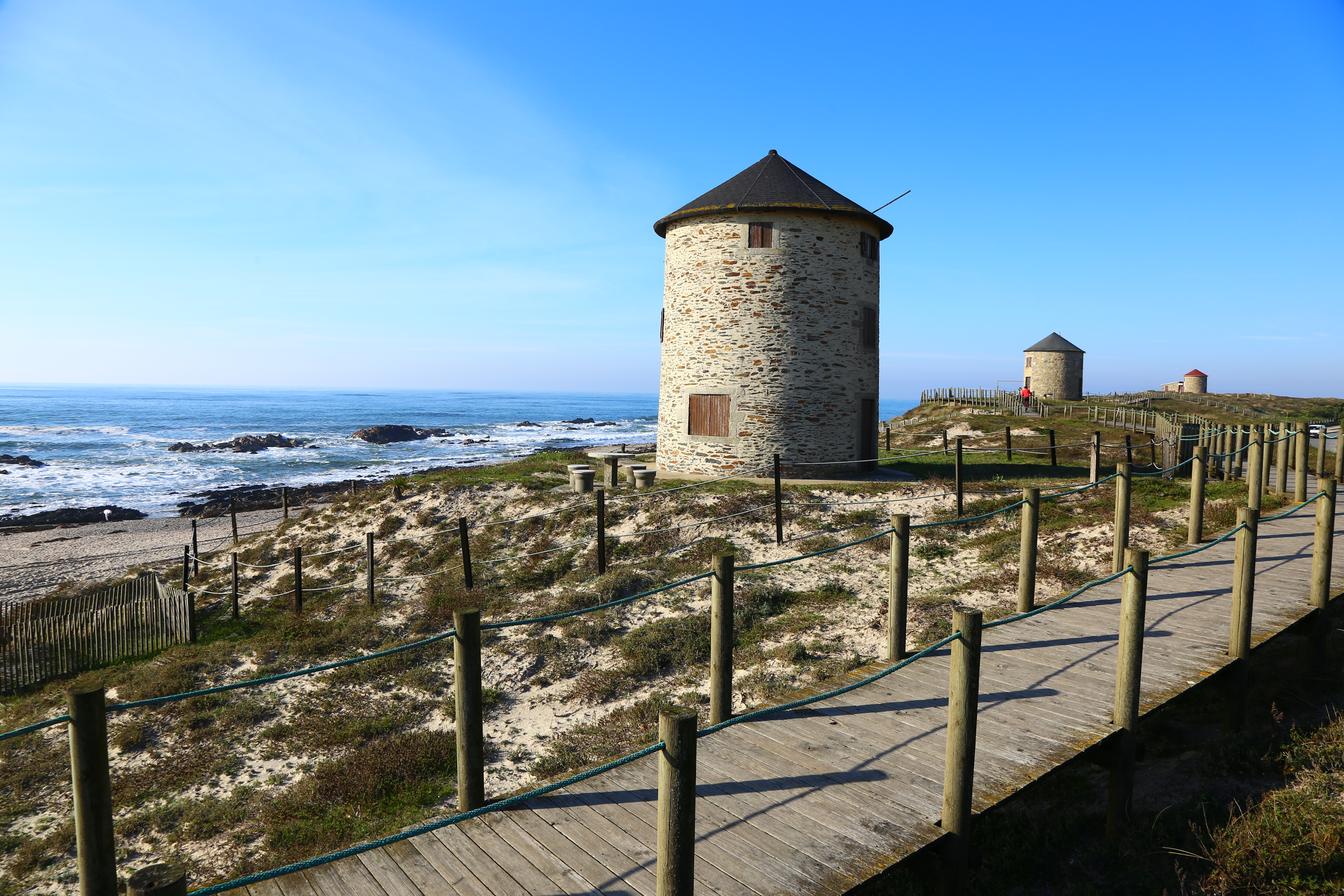 A cylindrical stone structure with a cone-shaped roof sits just off the ocean, where waves are crashing into the shore. An empty boardwalk is to the right, wrapping around the coast line.