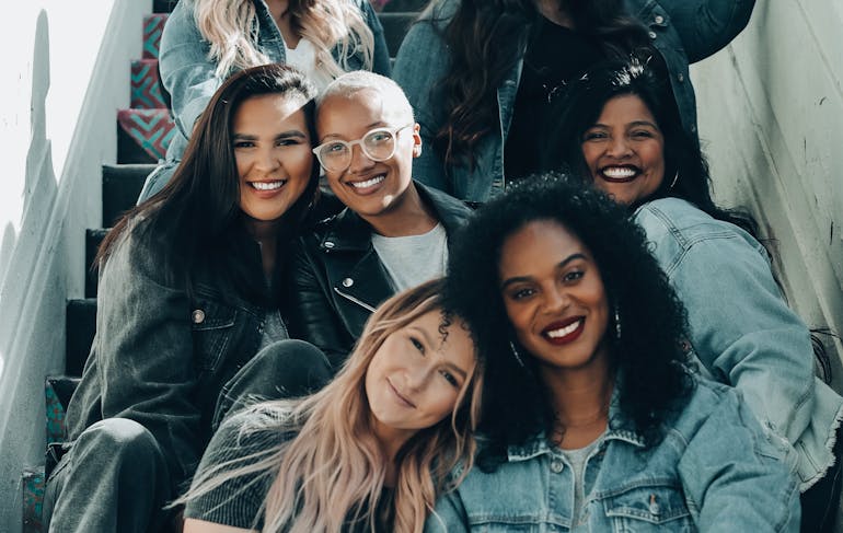 A group of 7 diverse women of all different skin tones sit on steps facing the camera and smiling.