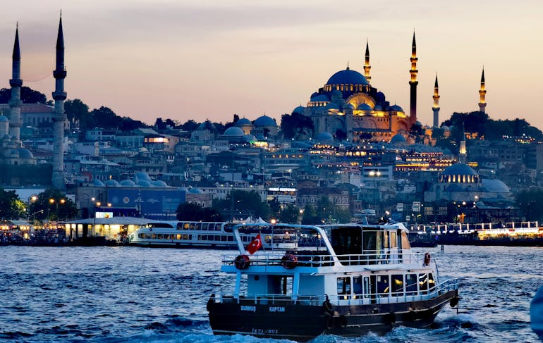 A boat is sitting in the harbor in front of Istanbul. The day is shifting to night as the city is lit with warm lights while there still is a warmly lit sky in the background. You can see the historic Hagia Sophia towering over the city and dominating the sky line. 