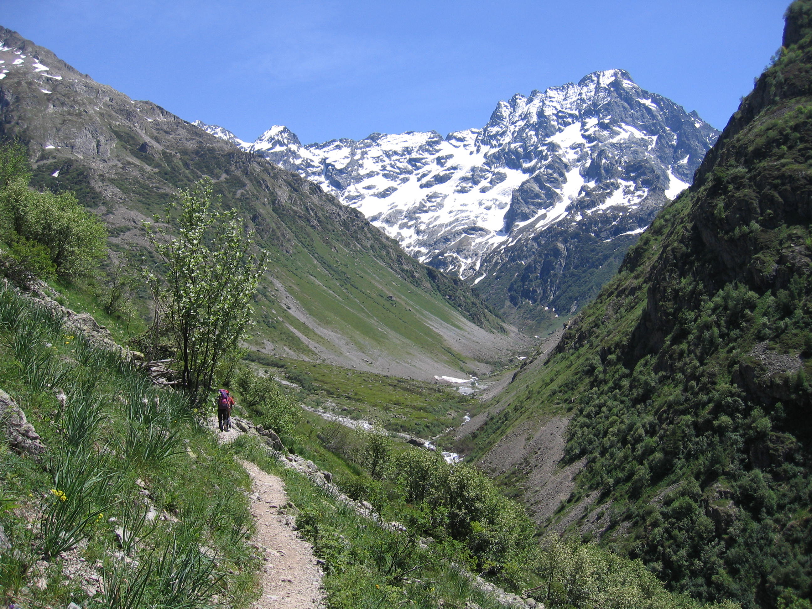 A hiker walks along a dirt path through a dramatic valley with steep green slopes on both sides and a prominent, jagged, snow-capped mountain peak in the background.