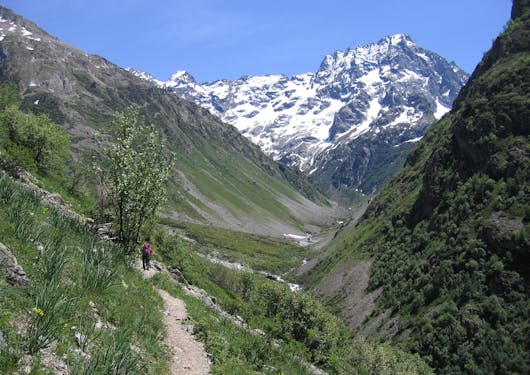 A hiker walks along a dirt path through a dramatic valley with steep green slopes on both sides and a prominent, jagged, snow-capped mountain peak in the background.