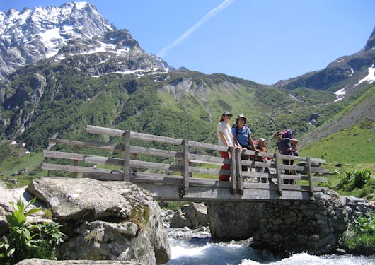 A group of 4 White travelers, two carrying day packs, stand on a wooden bridge over a rushing stream with greenery behind them and a French Alps peak in the background. It's a sunny blue sky day. Responsible package trips in France.