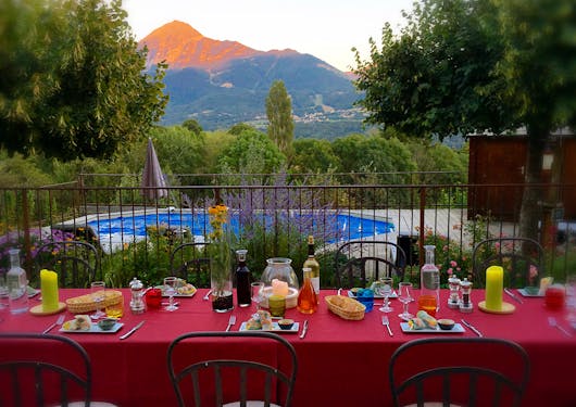 A lunch spread of freshly made spring rolls, bread, wine, and candles is on a bright red table cloth in front of a metal railing separating the eating area from a pool, with a peak of the southern French Alps in the distance with a golden sunset on its tip. Greenery is all around. Responsible package trips in France.