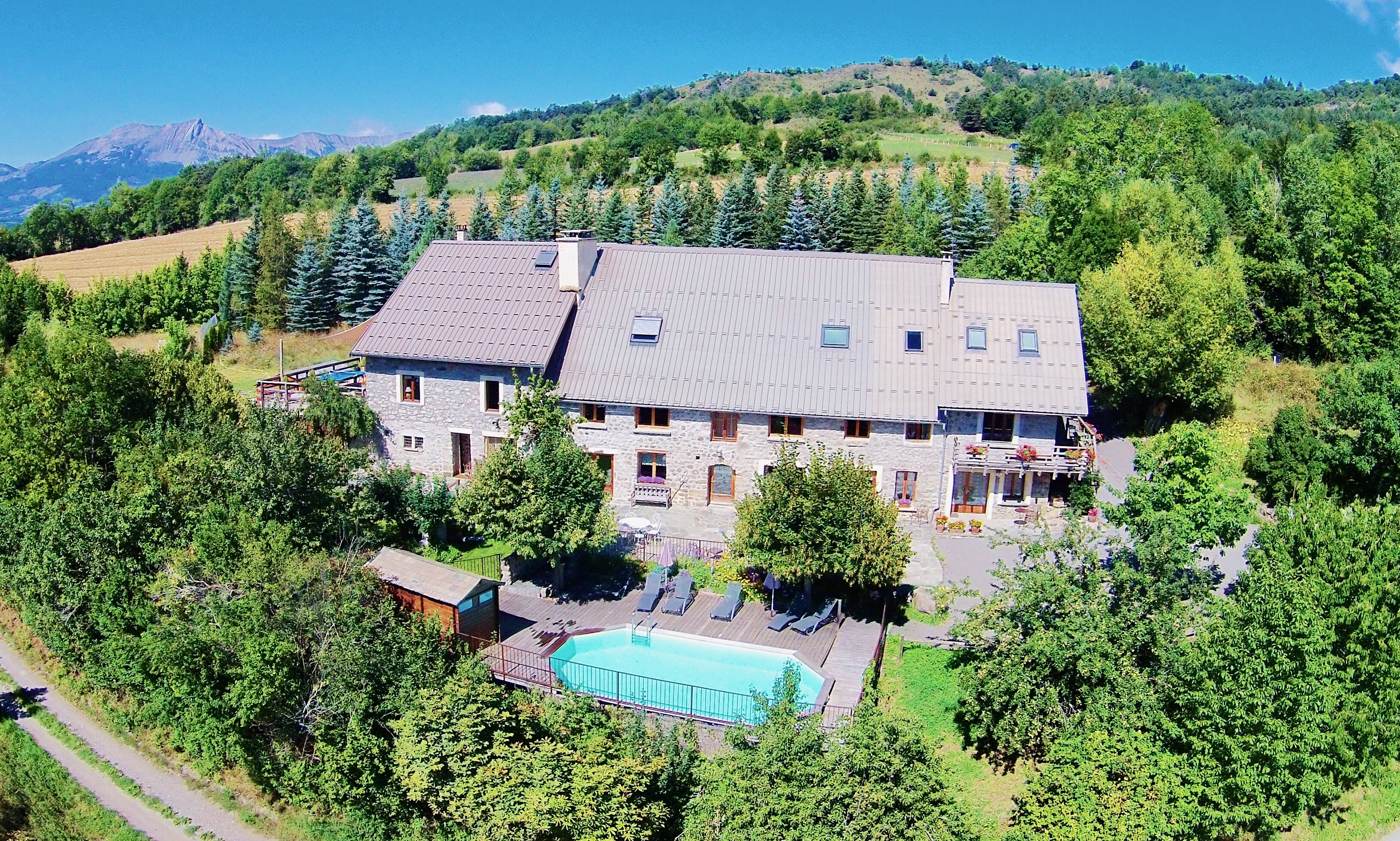 An alpine lodge is nestled amongst bright green trees, with a blue sky overhead and a teal pool in the foreground. It's an aerial shot showing the entire property. Responsible package trips in France.