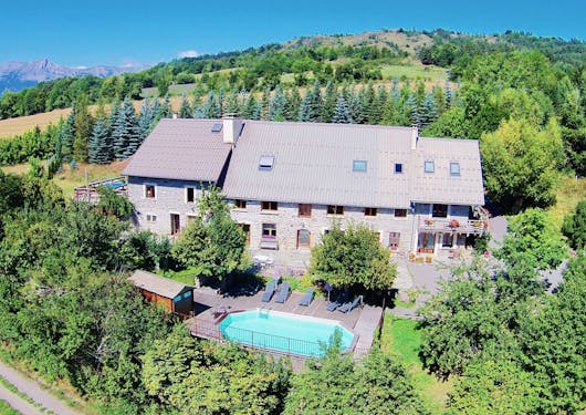 An alpine lodge is nestled amongst bright green trees, with a blue sky overhead and a teal pool in the foreground. It's an aerial shot showing the entire property. Responsible package trips in France.