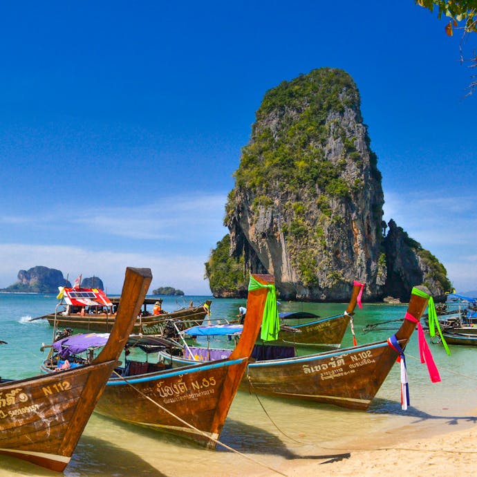 Traditional wooden boats in Thailand on the beach of Krabi are shown in shallow water with light blue water behind them and an impressive rock structure in the distance. Discover sustainable package trips in Thailand!