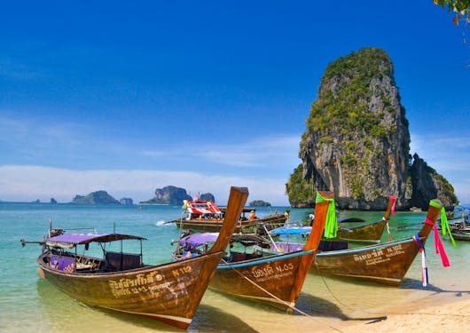 Traditional wooden boats in Thailand on the beach of Krabi are shown in shallow water with light blue water behind them and an impressive rock structure in the distance. Discover sustainable package trips in Thailand!