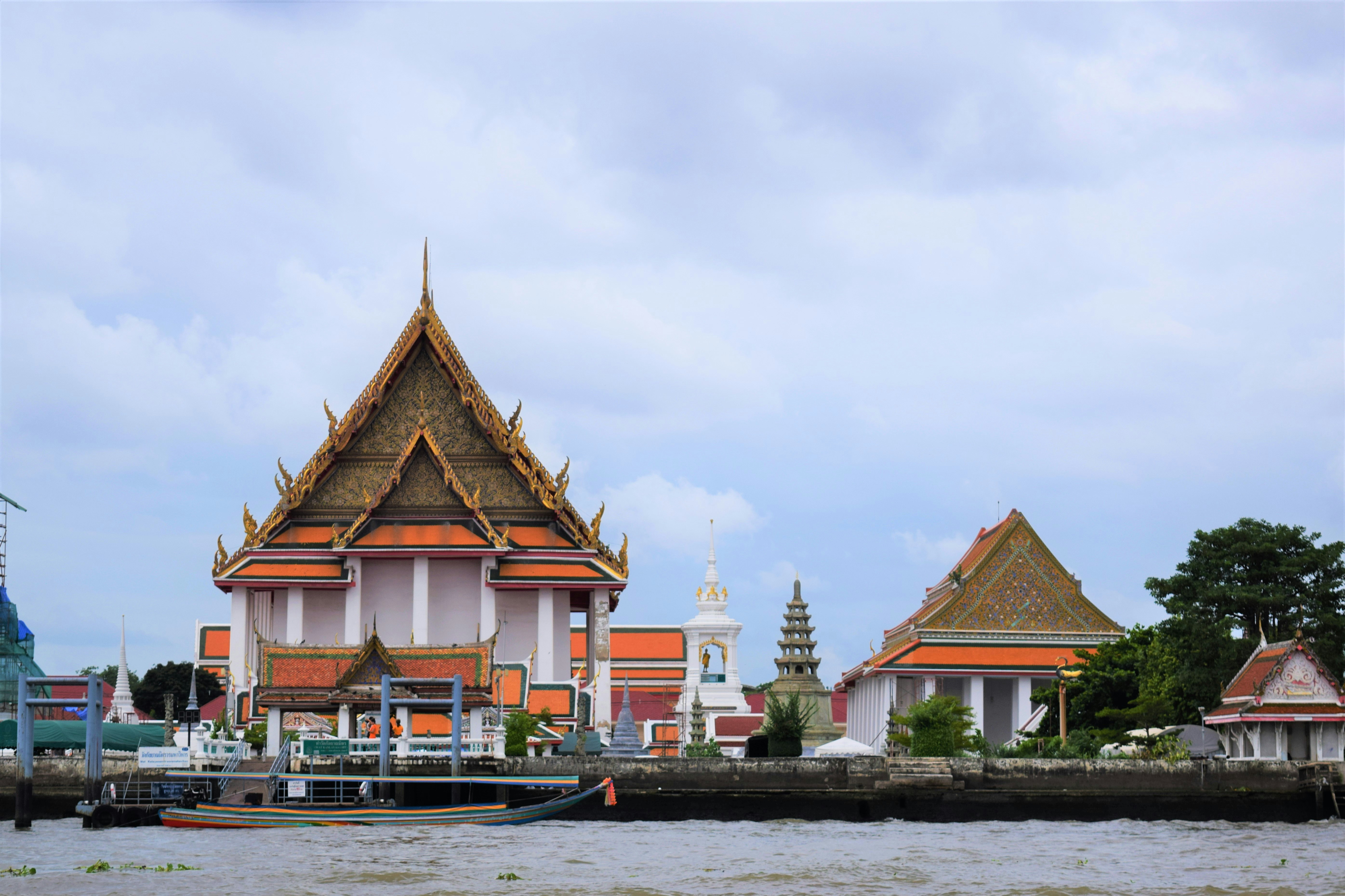 Temples and small structures are seem from the Chao Praya River in Bangkok. The cloud has a light sky cover.