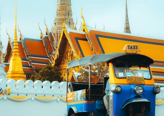 A blue and yellow tuktuk in Bangkok, Thailand is shown in from of a bright yellow and orange temple.