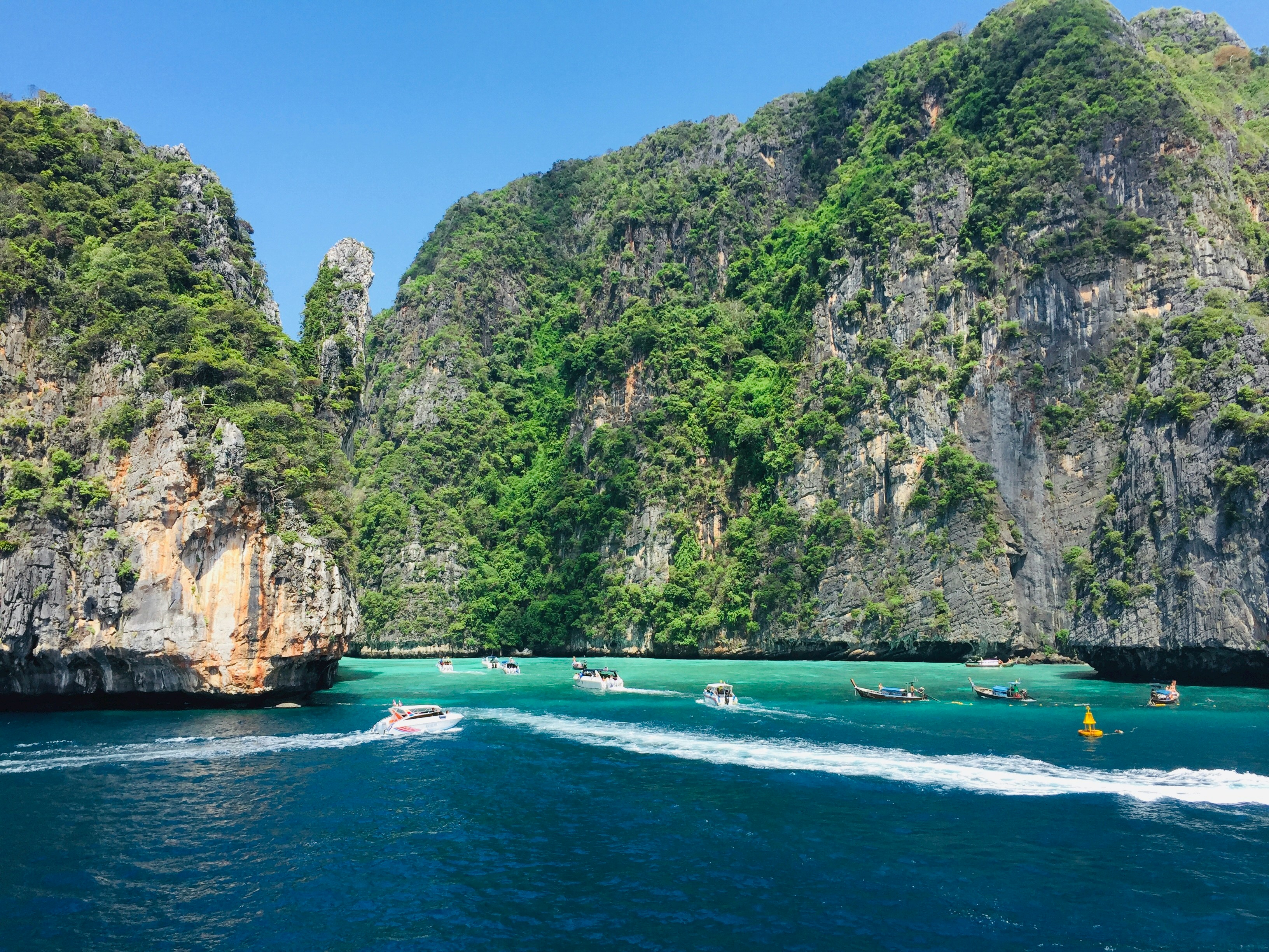 The stunning Phi Phi Islands in Thailand are shown with bright blue and teal water in the foreground and bright green trees going up the rocky cliffs of the islands. About 10 small boats are milling about between the islands. It's a sunny blue sky day.