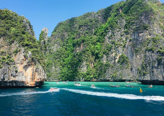 The stunning Phi Phi Islands in Thailand are shown with bright blue and teal water in the foreground and bright green trees going up the rocky cliffs of the islands. About 10 small boats are milling about between the islands. It's a sunny blue sky day.
