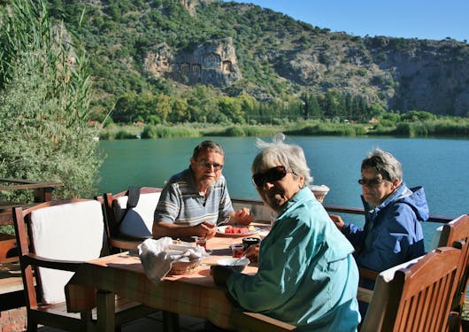 A group of 3 adults, Caucasians in their 50s or 60s, sit, smiling over lunch along the Lycian Way in Turkey with blue-green water in the background and an interesting rock formation built into the hill across the water.