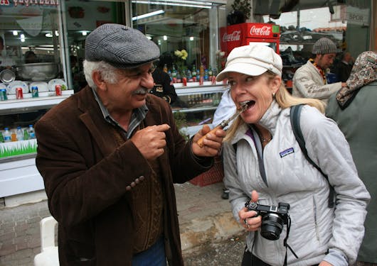 A local Turkish man, grinning and wearing a brown jacket with a grey plaid newsboy hat, feeds a bit of a local delicacy off the tip of a knife to a Caucasian, adult female traveler wearing a light grey jacket and white hat, and holding a black camera.