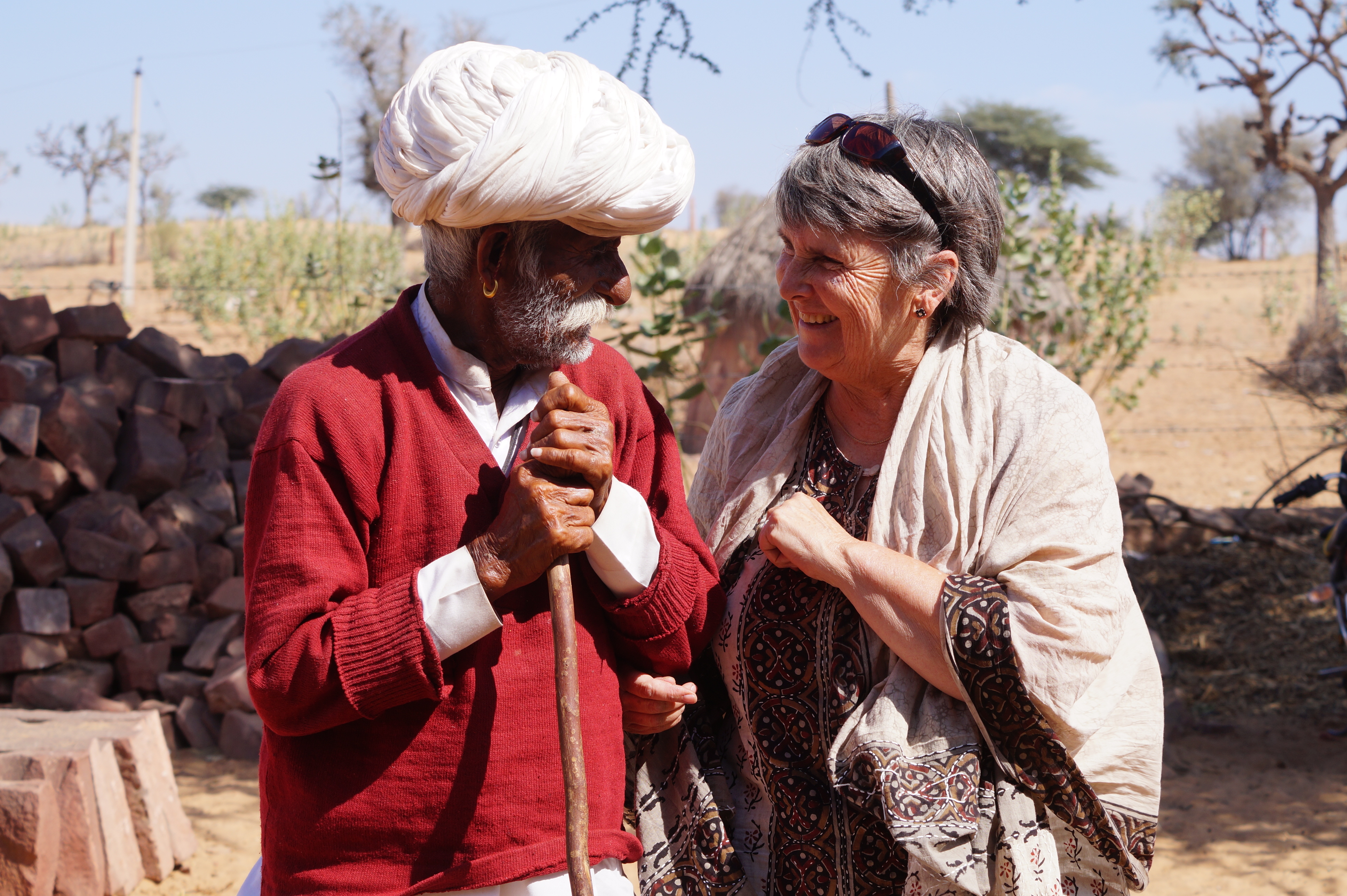 A Caucasian woman in her 60's or 70's with silver hair stands next to an Indian man, smiling. She's covered in a beige shawl while the Indian man has a white turban on and a long sleeve red sweater. 