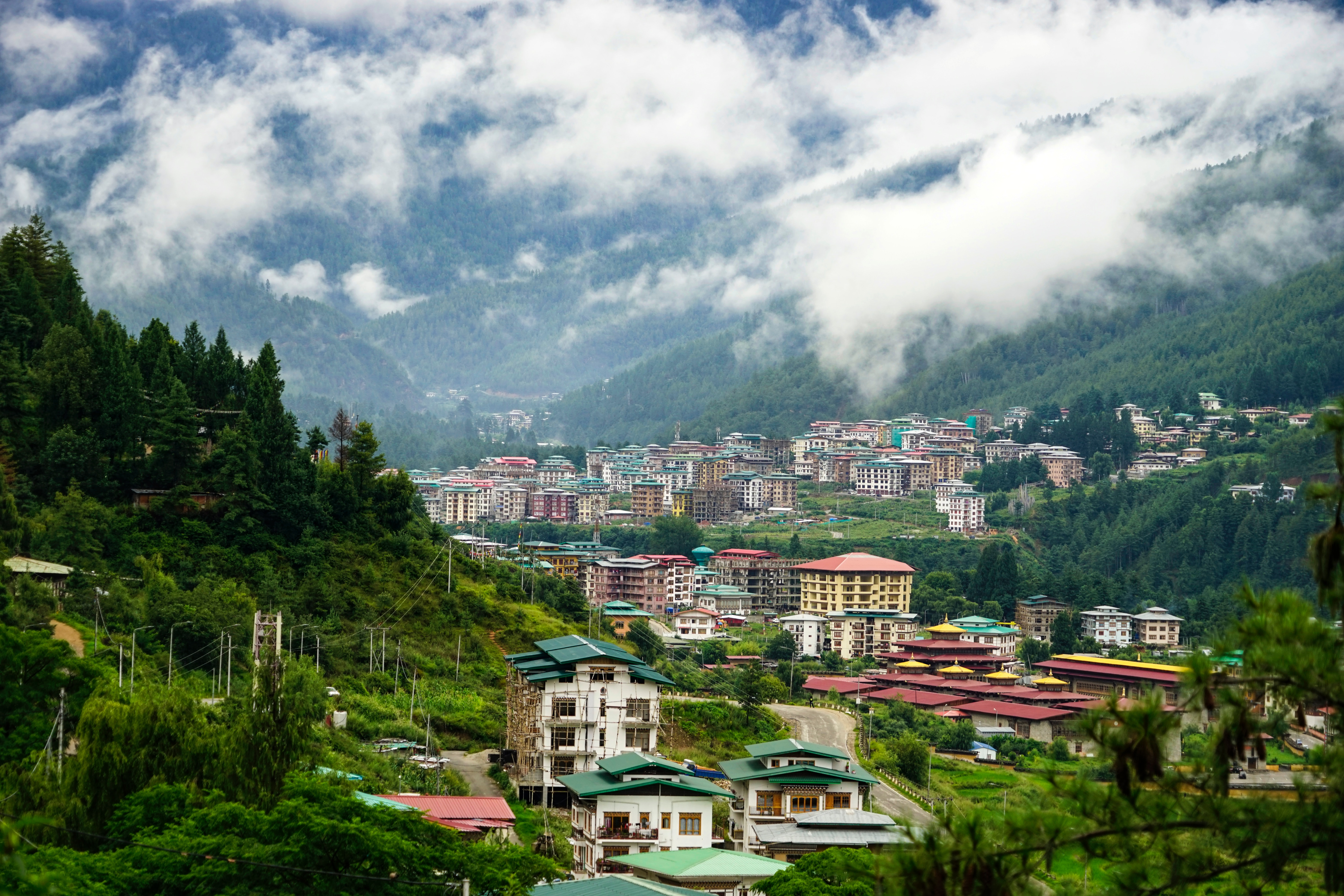 Colorful houses with green and red roofs sprawl along the Thimphu Valley in Bhutan. The Valley is lined with green trees and hills, and a cloudy fog is coming over the hills onto the city.